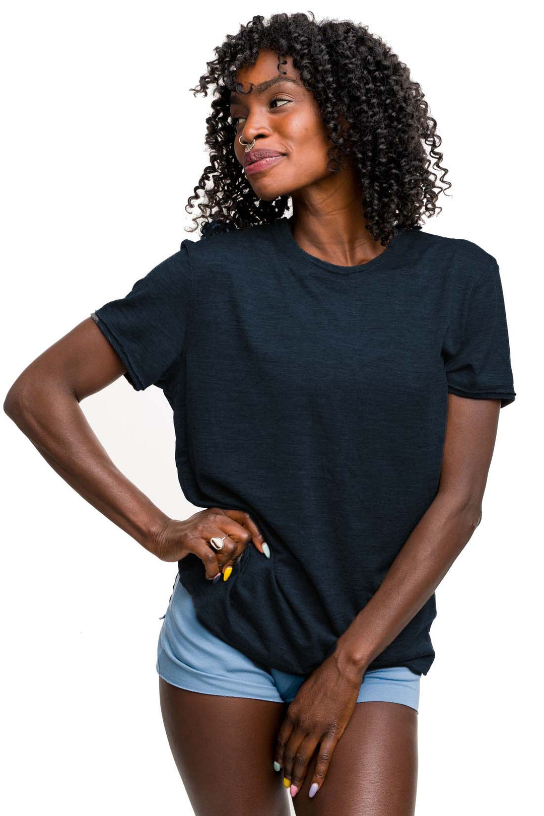 A woman with curly black hair wearing a dark navy blue short-sleeve merino wool crewneck t-shirt and light blue bamboo shorts, posing against a solid white background.