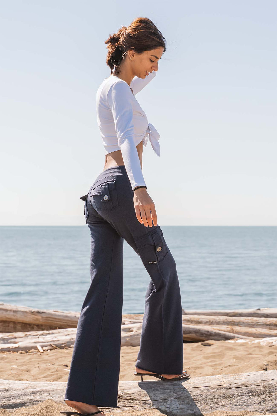 A woman stands on a sandy beach wearing dark navy wide-leg cargo pants and a white tied long-sleeve crop top, with the ocean and sky in the background.